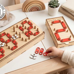 May include: A wooden puzzle set featuring two rectangular boxes with pegs and red and white connector pieces to form numbers. The set includes digital download files for laser cutting, with text indicating 2 designs and 3mm material width.