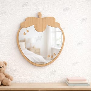 May include: A wooden strawberry-shaped mirror with a natural finish hangs on a white wall. The mirror has a clear reflective surface and teardrop cutouts. A teddy bear and stacked books sit below. The material width is 3mm.