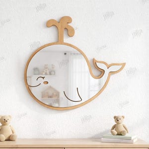 May include: A wooden whale-shaped mirror with a natural wood finish. The mirror has a round body, a tail, and a spout. The mirror is mounted on a white wall. The material width is 3mm.