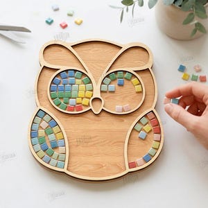 May include: Wooden owl-shaped mosaic tray with colorful square tiles. The tray has a light brown wood grain finish. The eyes and feathers of the owl are designed to hold the mosaic tiles. A hand is placing a tile.