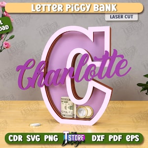 Puede incluir: Una hucha de madera con forma de letra "C" en color lila claro, con el nombre "Charlotte" en cursiva. El banco contiene dinero y monedas. Las palabras "Letter Piggy Bank" y "Laser Cut" son visibles.