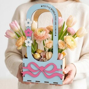 May include: A wooden floral arrangement with a light blue frame and a pink bow. The arrangement includes pink and yellow flowers, chocolates, and macarons. The text "TO THE WORLD YOU ARE A MOM" is on the frame.