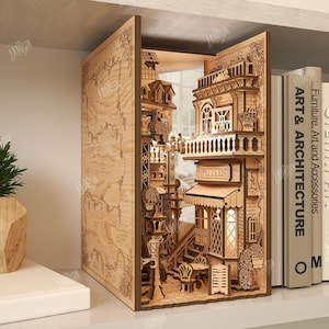 May include: A laser-cut wooden book nook featuring a detailed miniature street scene with buildings, stairs, and a cafe. The book nook is illuminated and stands on a shelf next to books and a potted plant. The material width is 3mm.