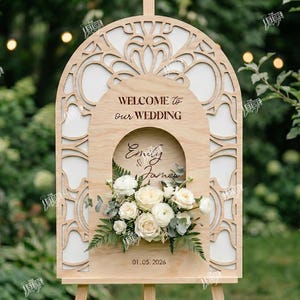 May include: A wooden wedding welcome sign with an arched top and floral accents. The sign reads "Welcome to our Wedding" and features the names "Emily & James" and the date "01.05.2026". The sign is supported by a wooden easel.