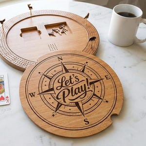 K&ouml;nnte beinhalten: Ein h&ouml;lzernes Cribbage-Brett mit Stiften, W&uuml;rfeln und Spielkarten. Das Brett hat ein kreisf&ouml;rmiges Design mit einer Windrose und den Worten "Let's Play!" auf dem Deckel eingraviert. Eine wei&szlig;e Tasse ist ebenfalls vorhanden.