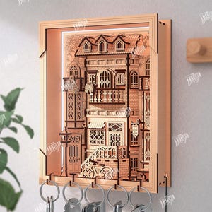 May include: A wooden key holder featuring a detailed laser-cut design of a building facade. The key holder has a rectangular frame and includes key hooks at the bottom. The text "THE STORE DESIGN" and "CUT READY FILE DIGITAL DOWNLOAD" are visible. The material width is 3mm.
