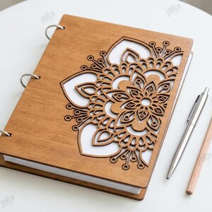 May include: A wooden notebook with a laser-cut floral design on the cover. The notebook is open, revealing white pages. A silver pen and a pencil are placed beside the notebook. The notebook is a digital download.