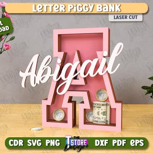 Puede incluir: Hucha con forma de letra "A" rosa con el nombre "Abigail" en escritura blanca. El banco contiene monedas y un billete. Las palabras "Letter Piggy Bank" y "Laser Cut" son visibles.