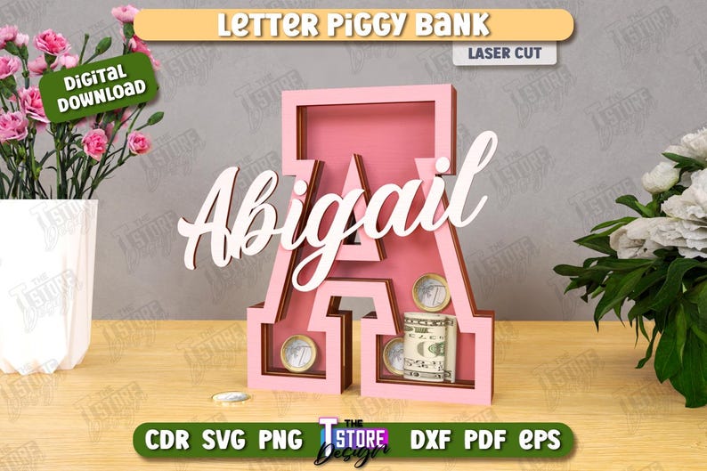Puede incluir: Hucha con forma de letra "A" rosa con el nombre "Abigail" en escritura blanca. El banco contiene monedas y un billete. Las palabras "Letter Piggy Bank" y "Laser Cut" son visibles.