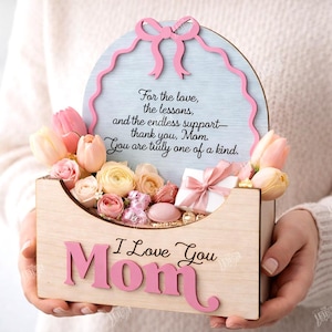 May include: A wooden Mother's Day gift box with the words "I Love You Mom" in pink. The box contains flowers and a small gift. A light blue sign with a pink border and a message is behind the flowers.