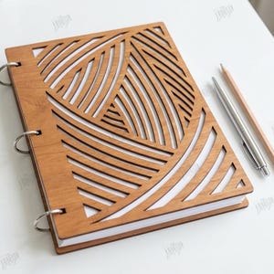 May include: A wooden notebook with a geometric design cut into the cover. The cover is light brown and has a pattern of curved lines. The notebook is open, revealing white pages. A pen and pencil are next to the notebook.