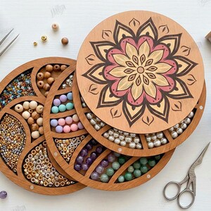 May include: Wooden bead organizer with a floral design on the lid. The organizer is round with multiple compartments filled with various beads in different colors and sizes. A pair of tweezers and small scissors are also visible.