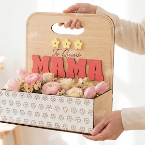 May include: Wooden flower box with the word "MAMA" in red letters and the phrase "Te Quiero" above. The box is filled with pink and white flowers. The box has a handle and a floral pattern.