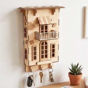 May include: A light-colored wooden wall-mounted key holder shaped like a building facade with a balcony, awning, and windows. It features three metal hooks at the bottom for hanging keys. The design includes laser-cut details and a 3mm material width.