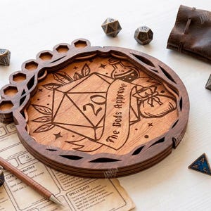 May include: A wooden dice tray with a carved design featuring a D20 die, potion bottles, and a banner that reads "The Gods Approve." The tray has a circular shape with a dice holder and is surrounded by several dice and a leather pouch.