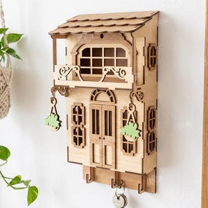 May include: A laser-cut wooden wall-mounted key holder shaped like a two-story house with a balcony, windows, and hanging plant details. The structure features a shingled roof, a central door, and three hooks at the bottom for keys. Material width: 3mm.