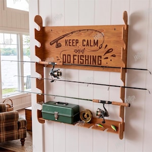 May include: Wooden fishing rod rack with the text "KEEP CALM and DO FISHING". The rack holds three fishing rods, a green tackle box, and fishing lures. The rack is mounted on a white wall.
