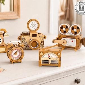 Puede incluir: Una colección de cinco diseños de corte láser de madera, que incluyen un teléfono, una cámara, un despertador, una radio y una cámara de cine. Los diseños son de madera marrón claro y se muestran sobre una superficie blanca. El ancho del material es de 3 mm.