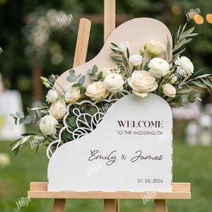 May include: A white welcome sign for a wedding, shaped like a piano, with floral arrangements and the names "Emily & James" and the date "01.05.2026". The sign is on a wooden easel.