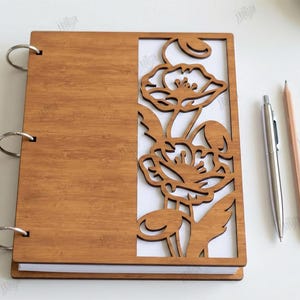 May include: A wooden notebook with a laser-cut floral design on the cover. The notebook is held together with silver rings. A silver pen and a pencil are next to the notebook. The text on the right says "CUT READY FILE DIGITAL DOWNLOAD".