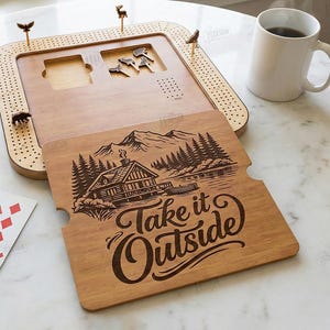 K&ouml;nnte beinhalten: Cribbage-Brett aus Holz mit Stiften und einem dekorativen Holzdeckel mit einer H&uuml;ttenlandschaft und dem Text "Take it Outside". Enth&auml;lt W&uuml;rfel und Spielkarten. Eine wei&szlig;e Tasse ist ebenfalls vorhanden.