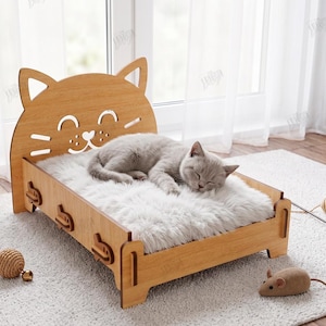 May include: A light brown wooden pet bed with a cat-shaped headboard featuring cut-out eyes, whiskers, and a nose. The bed includes a white faux fur cushion and is designed for a 12mm material width. The text reads The Store Design Cut Ready File.