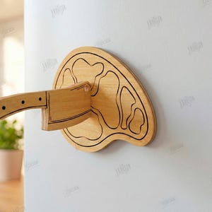Puede incluir: Una cuchilla de madera y una tabla de cortar de madera est&aacute;n montadas en una pared blanca. La cuchilla es de madera marr&oacute;n claro con detalles en negro. La tabla de cortar tiene una forma &uacute;nica y un contorno negro. Ancho del material: 3 mm.