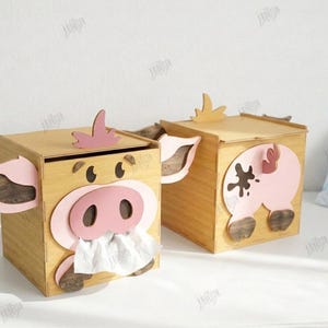 May include: Two wooden tissue box covers shaped like pigs. Each box has a light brown finish with pink accents for the ears, snout, and tail. One pig has a tissue sticking out of its snout. The boxes are 3mm wide.