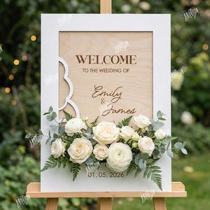 May include: A wooden wedding welcome sign with the text "WELCOME TO THE WEDDING OF Emily & James" and the date 01.05.2026. The sign is framed in white and decorated with white roses and greenery. The sign is on an easel.