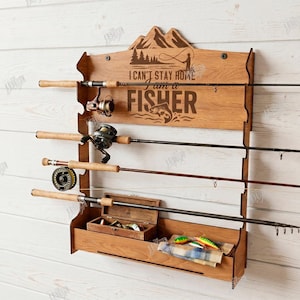 May include: Wooden fishing rod rack with the text "I CAN'T STAY HOME I AM A FISHER". The rack has a mountain and fishing scene design. It holds four fishing rods, a tackle box, and fishing lures. The rack is mounted on a white wooden wall.