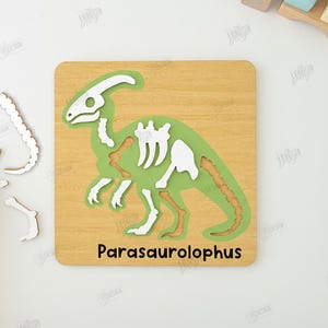 K&ouml;nnte beinhalten: Ein Holzpuzzle mit einem gr&uuml;nen Parasaurolophus-Dinosaurier mit wei&szlig;en und braunen Knochen. Das Puzzle befindet sich auf einer hellbraunen Holzbasis, auf der der Name des Dinosauriers aufgedruckt ist. Einige Puzzleteile sind separat.