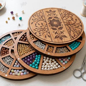 May include: Wooden bead organizer with multiple compartments filled with colorful beads. The organizer has a floral design on the lid. A small plate with a bracelet, tweezers, and scissors are also visible.