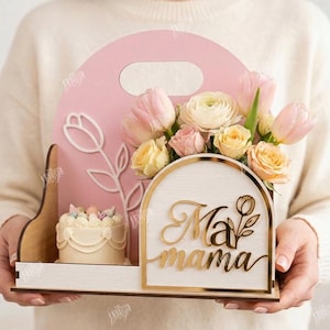 May include: A pink wooden Mother's Day gift box with a handle, holding a floral arrangement of pink and yellow flowers, a small cake, and the words "Ma mama" in gold script. The box has a tulip design.