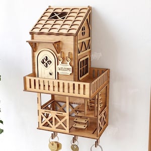 May include: A light brown wooden wall-mounted key holder shaped like a two-story house with a balcony, a door with the text WELCOME, and three metal hooks at the bottom. The structure features laser-cut details, a tiled roof, and a staircase.