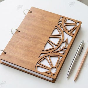 May include: A wooden notebook with a laser-cut design of citrus fruit and leaves. The notebook is open, revealing white pages. A silver pen and a wooden pencil are placed next to the notebook. The image also includes the text "CUT READY FILE DIGITAL DOWNLOAD".