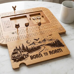 K&ouml;nnte beinhalten: Ein h&ouml;lzernes Cribbage-Brett mit passendem Deckel mit einem Roadtrip-Design und dem Text "BORN TO ROAM". Das Brett enth&auml;lt tierf&ouml;rmige Stifte und einen Stauraum. Eine wei&szlig;e Tasse und W&uuml;rfel sind ebenfalls sichtbar.