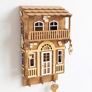 May include: A laser-cut wooden wall-mounted key holder shaped like a two-story house with a balcony, white window frames, and a white door. The structure features two hanging lanterns, three hooks at the bottom, and a 3mm material width label.