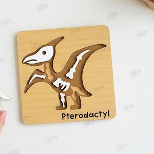 K&ouml;nnte beinhalten: Holzpuzzle Pterodactyl mit Dinosaurier-Skelett-Design. Das Puzzle ist hellbraun mit wei&szlig;en und braunen Details. Das Wort "Pterodactyl" ist unten aufgedruckt. Enth&auml;lt schnittfertige Dateien.