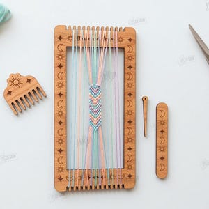 Op de afbeelding: Een houten weefgetouw met een geweven armband in pastelkleuren, een kam en andere gereedschappen. Het weefgetouw is versierd met zon- en maansymbolen. Borduurgaren in pastelkleuren is ook aanwezig.