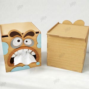 May include: Two wooden tissue box holders. One features a cartoon face with an open mouth and tissue paper. The other is a plain wooden box. The boxes are made of light brown wood with blue and gray accents. The image also includes a stuffed bunny.