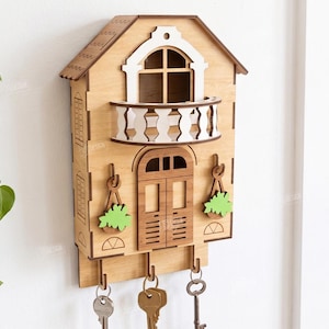 May include: A light brown wooden wall-mounted key holder shaped like a house with a white balcony, a central door, and two decorative hanging plant cutouts. Three metal hooks extend from the bottom, each holding a metal key. Text includes 3MM.