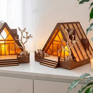 Può includere: Due modelli di case in legno a forma di A con interni illuminati, dotati di terrazza anteriore, scale e un ritaglio decorativo a forma di albero. Le strutture sono realizzate in materiale di 3mm di spessore, progettate come file di taglio laser in formati CDR, SVG, PNG, DXF, PDF ed EPS.