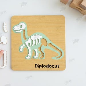 K&ouml;nnte beinhalten: Holz-Dinosaurier-Puzzle mit einem hellgr&uuml;nen Diplodocus-Skelett auf einer hellbraunen quadratischen Tafel. Das Puzzle enth&auml;lt separate wei&szlig;e Teile. Der Text "Diplodocus" steht unten. Kompatibel mit jeder Materialst&auml;rke.
