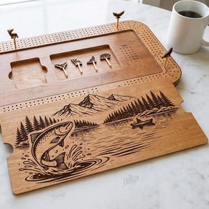 K&ouml;nnte beinhalten: Cribbage-Brett aus Holz mit einem lasergravierten Design eines Fisches, der aus dem Wasser springt, Berge und B&auml;ume. Enth&auml;lt Stifte in Tierform. Das Brett wird von Spielkarten, W&uuml;rfeln und einer Kaffeetasse begleitet.
