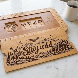 K&ouml;nnte beinhalten: Cribbage-Brett aus Holz mit einem "Stay Wild" Design. Das Brett zeigt eine detaillierte Berg- und Wildtierszene, darunter ein Hirsch, ein B&auml;r und ein Wolf. Enth&auml;lt Stifte und einen separaten Deckel. Eine Tasse Kaffee, W&uuml;rfel und eine Spielkarte sind ebenfalls sichtbar.