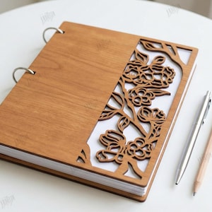 May include: A wooden notebook with a floral cut-out design on the cover. The notebook is held together with silver rings. A silver pen and a wooden pencil are placed beside the notebook. The text "CUT READY FILE DIGITAL DOWNLOAD" is visible.