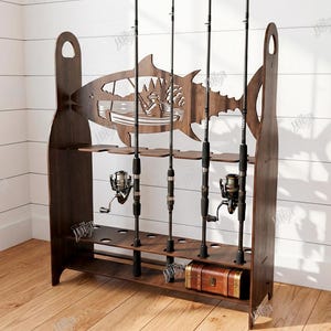 May include: A dark brown wooden fishing rod rack with a fish-shaped cutout design. The rack holds multiple fishing rods and reels, with a small wooden chest at the bottom. The design includes a silhouette of a person fishing in a boat.