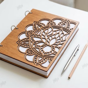 May include: A wooden notebook with a laser-cut floral design on the cover. The notebook is open, revealing white pages. A silver pen and a wooden pencil are placed beside the notebook. The notebook is held together with metal rings.