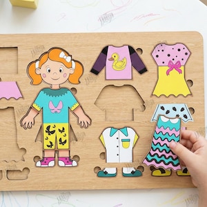 May include: Wooden puzzle with a girl figure and clothing pieces. Includes a teal top, pink skirt, yellow dress with a pink bow, and a blue and pink dress. The puzzle is compatible with any material thickness. The image also includes the text "CUT READY FILE".
