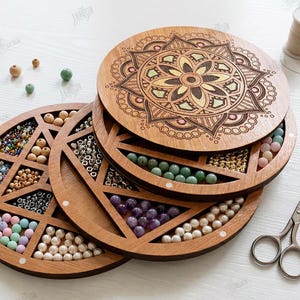 May include: Wooden bead organizer with a mandala design on the lid. The organizer is round with multiple compartments filled with colorful beads. A pair of small scissors and tweezers are also visible.